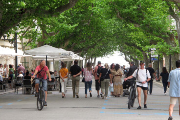 La voz de la comunidad sorda en Dénia: ¿Qué pasos se están dando para garantizar la plena accesibilidad? 3 Personas paseando y circulando en bicicleta por una calle peatonal arbolada de Dénia con terrazas a ambos lados.