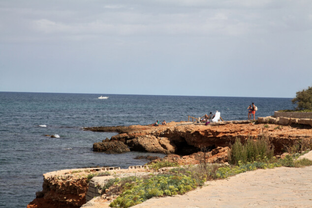 ¿Qué supone para Dénia la llegada del alga que arrasa con la pesca? 1 Imagen: Costa rocosa de Les Rotes, en Dénia, con varias personas junto al mar y una embarcación al fondo.