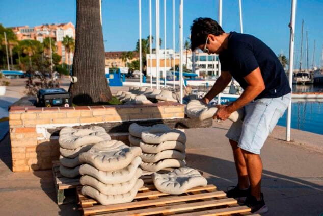 Imagen: Un hombre levanta una pieza modular de hormigón texturizado de una pila sobre un palé de madera en un puerto deportivo.