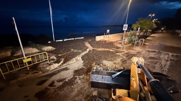 Dénia hace balance de la DANA: el Castillo y el polideportivo municipal cerrados y alerta en las calles 1 Imagen: Una pala cargadora amarilla retira de noche una gran cantidad de algas y escombros de un paseo marítimo inundado junto al mar.