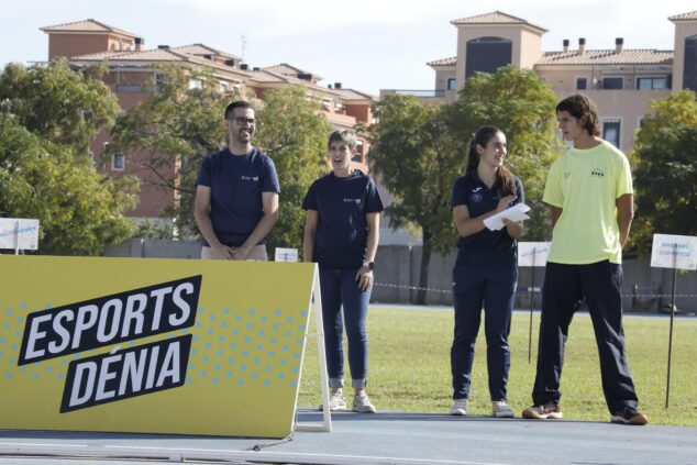 Imagen: Cuatro personas de pie junto a un cartel amarillo que dice 'ESPORTS DÉNIA' en una pista de atletismo durante un evento al aire libre con edificios al fondo.